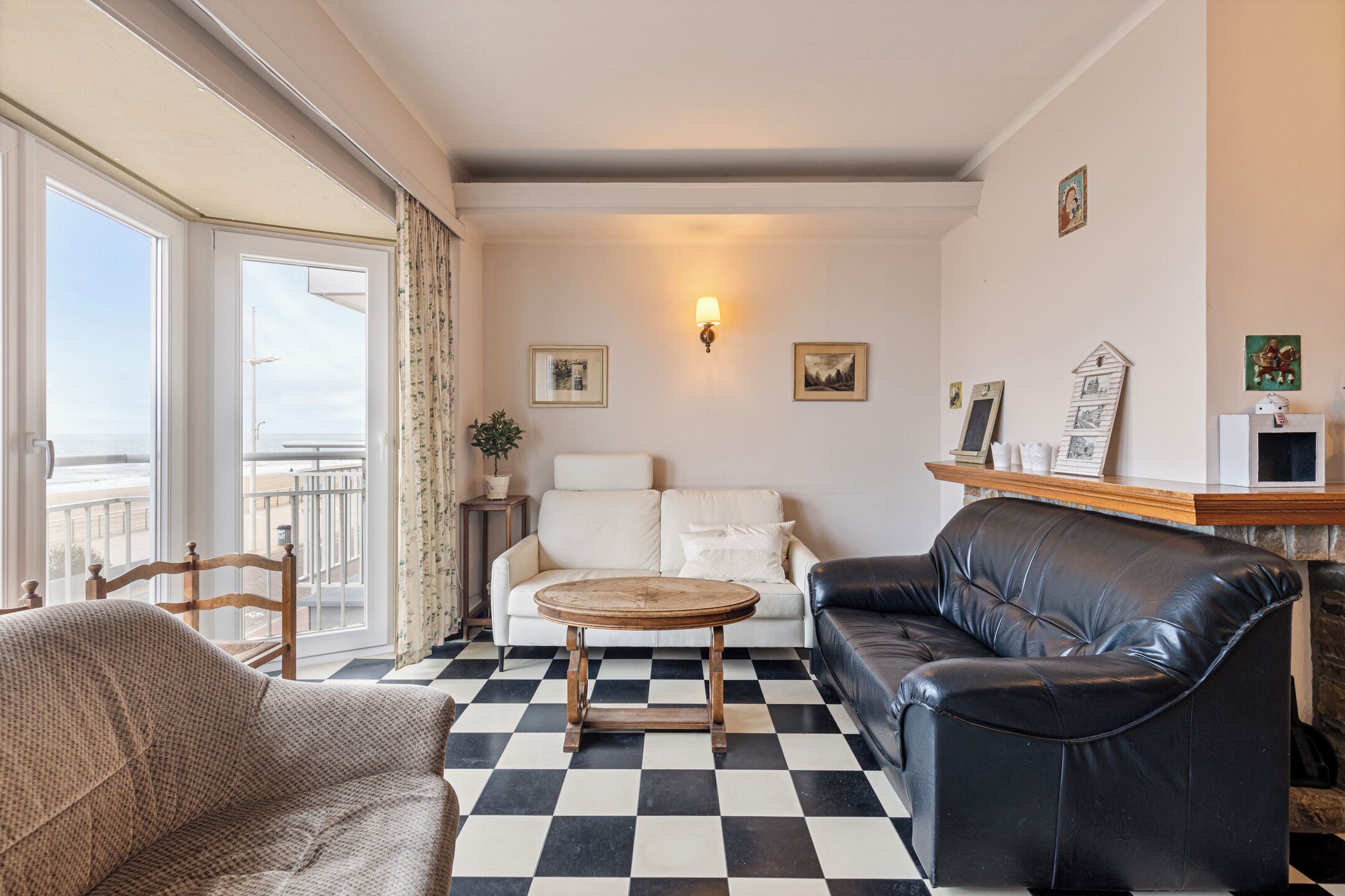 A living room with a checkered floor, a sofa, a chair, a round wooden table, and a balcony with a view.