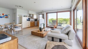 A modern living room with a sofa, coffee table, and dining area. There is a kitchen in the background with a large window. - Zonnehaven II E 0209+G47* (NIEUWPOORT)