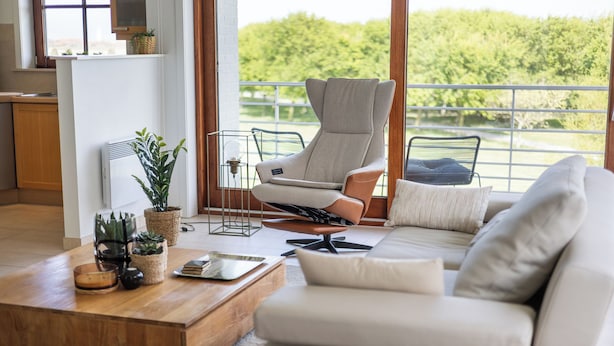 A modern living room with a sofa, a high-back chair, a coffee table, and a view of greenery through large windows. - Zonnehaven II E 0209+G47* (NIEUWPOORT)