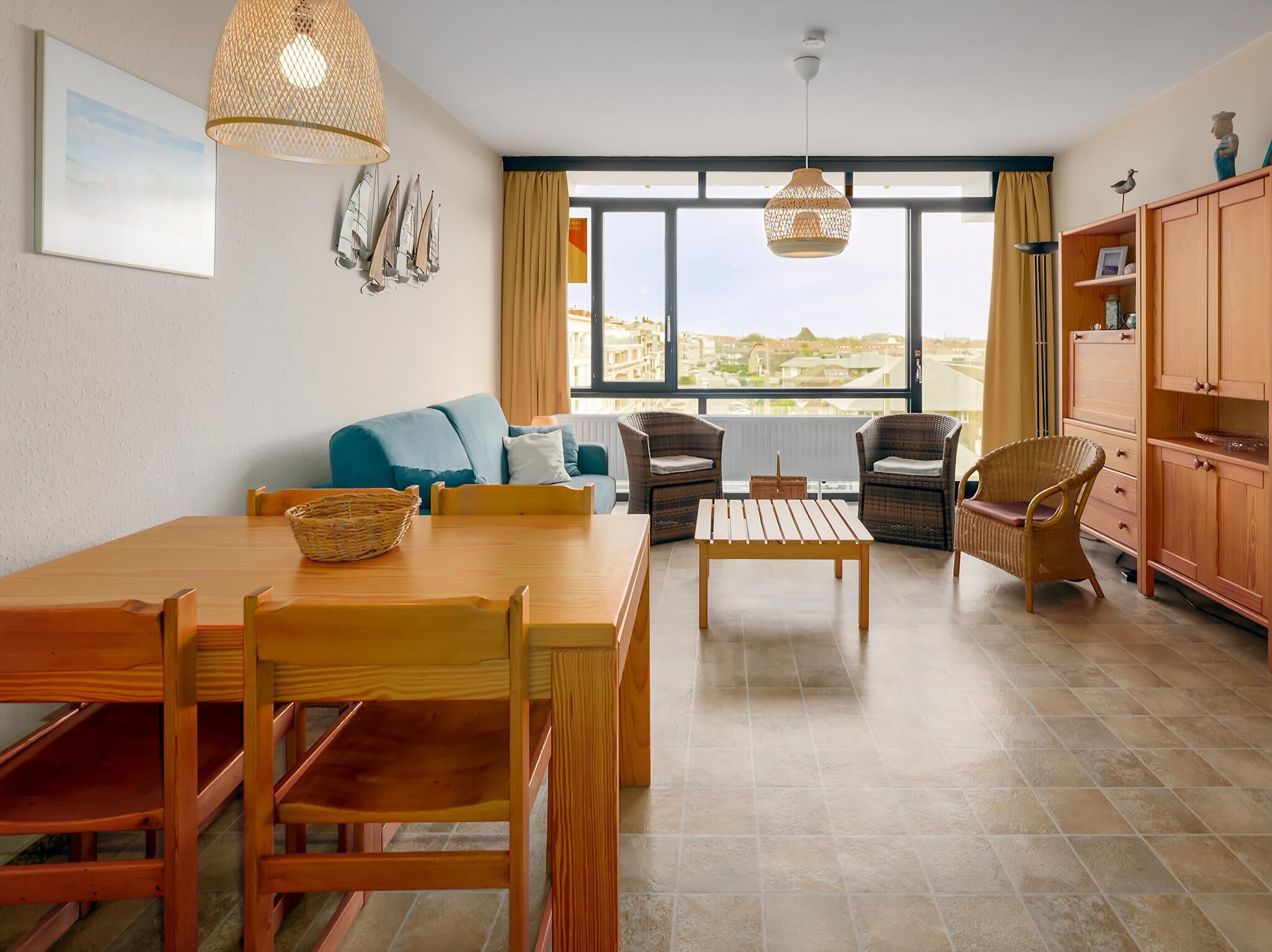 A spacious living area with a wooden dining table, wicker chairs, a blue sofa, and wicker armchairs. There are two hanging wicker lamps, a view through large windows, and a wooden cabinet with a shelf.