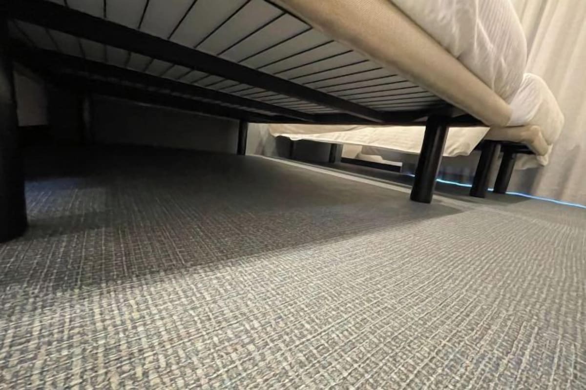 A carpeted floor with a bed frame underneath.