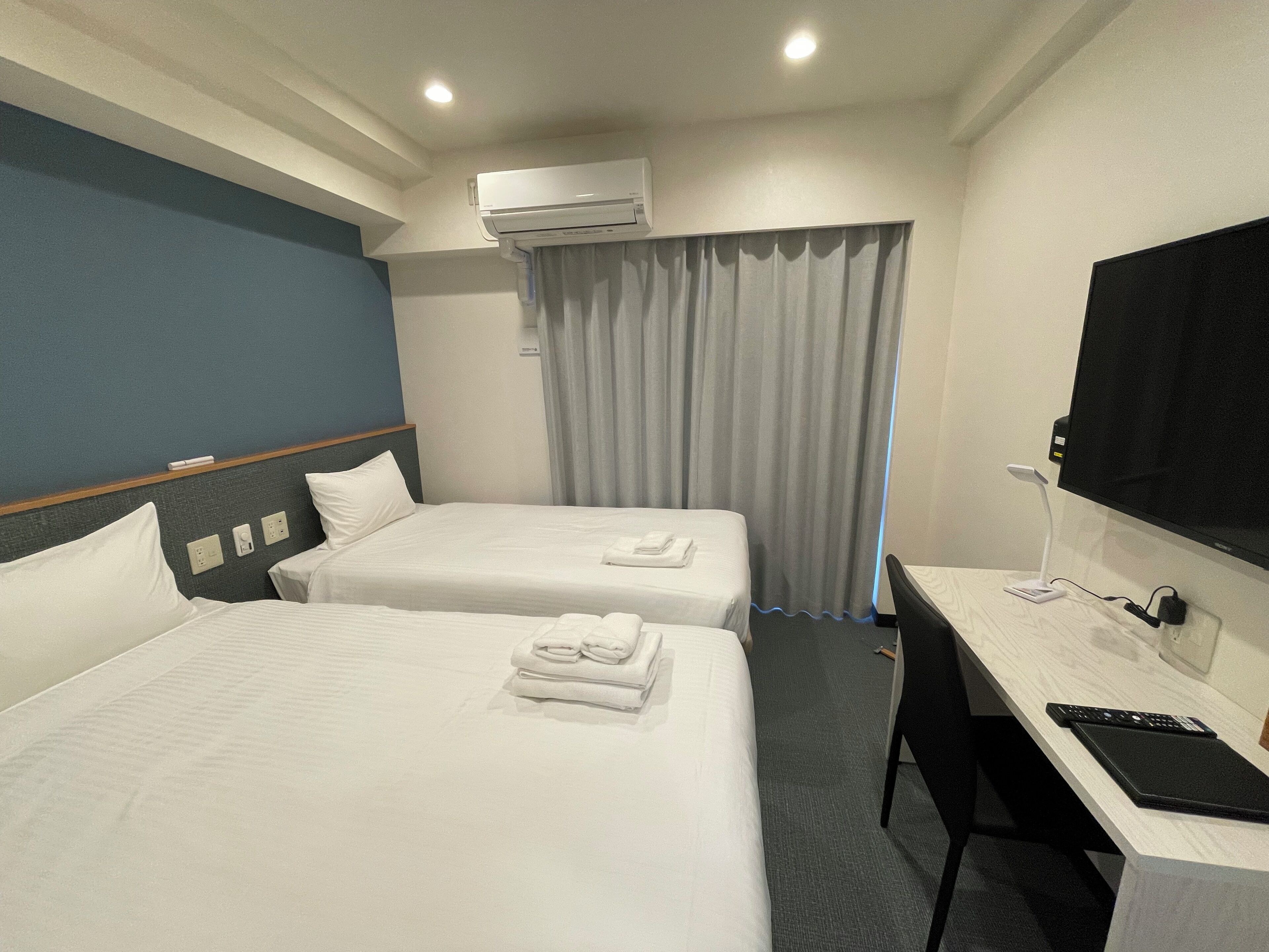 A hotel room with two beds, a desk, a TV, and an air conditioning unit.