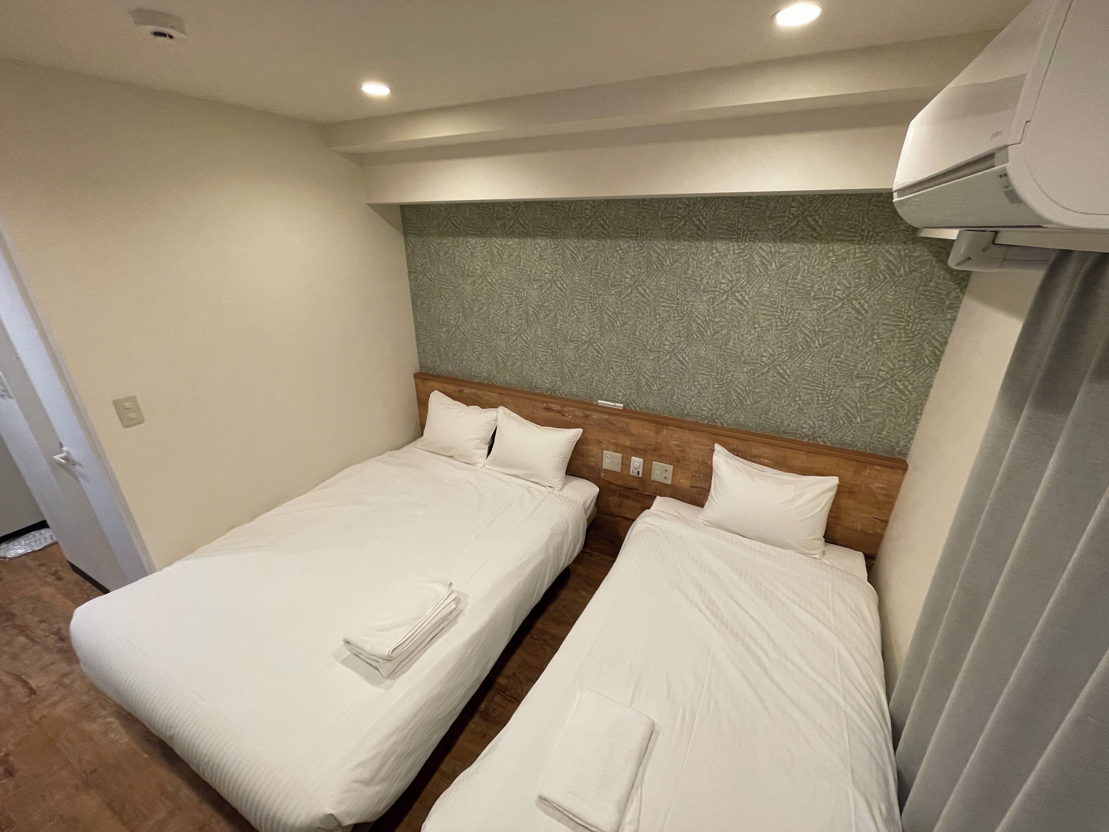 Habitación de hotel con dos camas individuales, cabecero de madera, pared texturizada de color verde y aire acondicionado instalado en el techo.