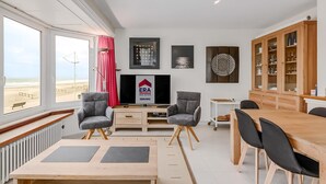 Una sala de estar moderna con un ventanal grande, un televisor de pantalla plana, una mesa de comedor de madera y un mueble con puertas de cristal.