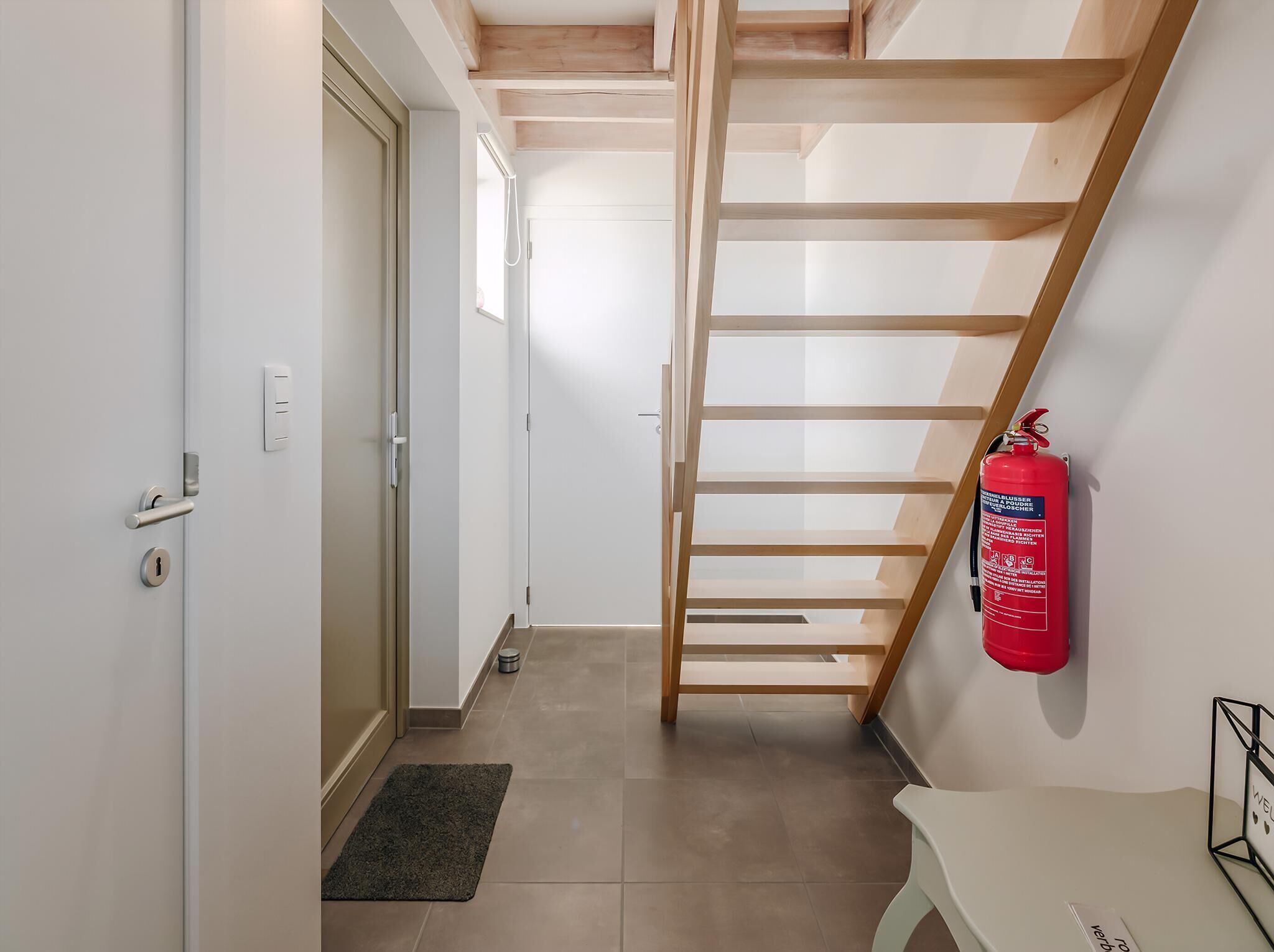 Un couloir étroit avec un escalier en bois, un extincteur et une petite table sur laquelle est posé un panneau.