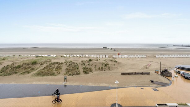 Une vue sur la plage avec un cycliste sur une piste pavée, des dunes de sable et une jetée au loin.