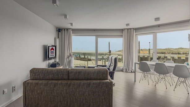A modern living room with a sofa, a dining area with chairs, and large windows offering a view of the cityscape. - Den Oever V 0106 met parking (Nieuwpoort)