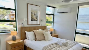 A bedroom with a bed, two bedside tables, a nightstand, a framed picture, and an air conditioning unit. - Headlands Beachside Gem - 50mtrs To Toes In Sand! (Alexandra Headland)