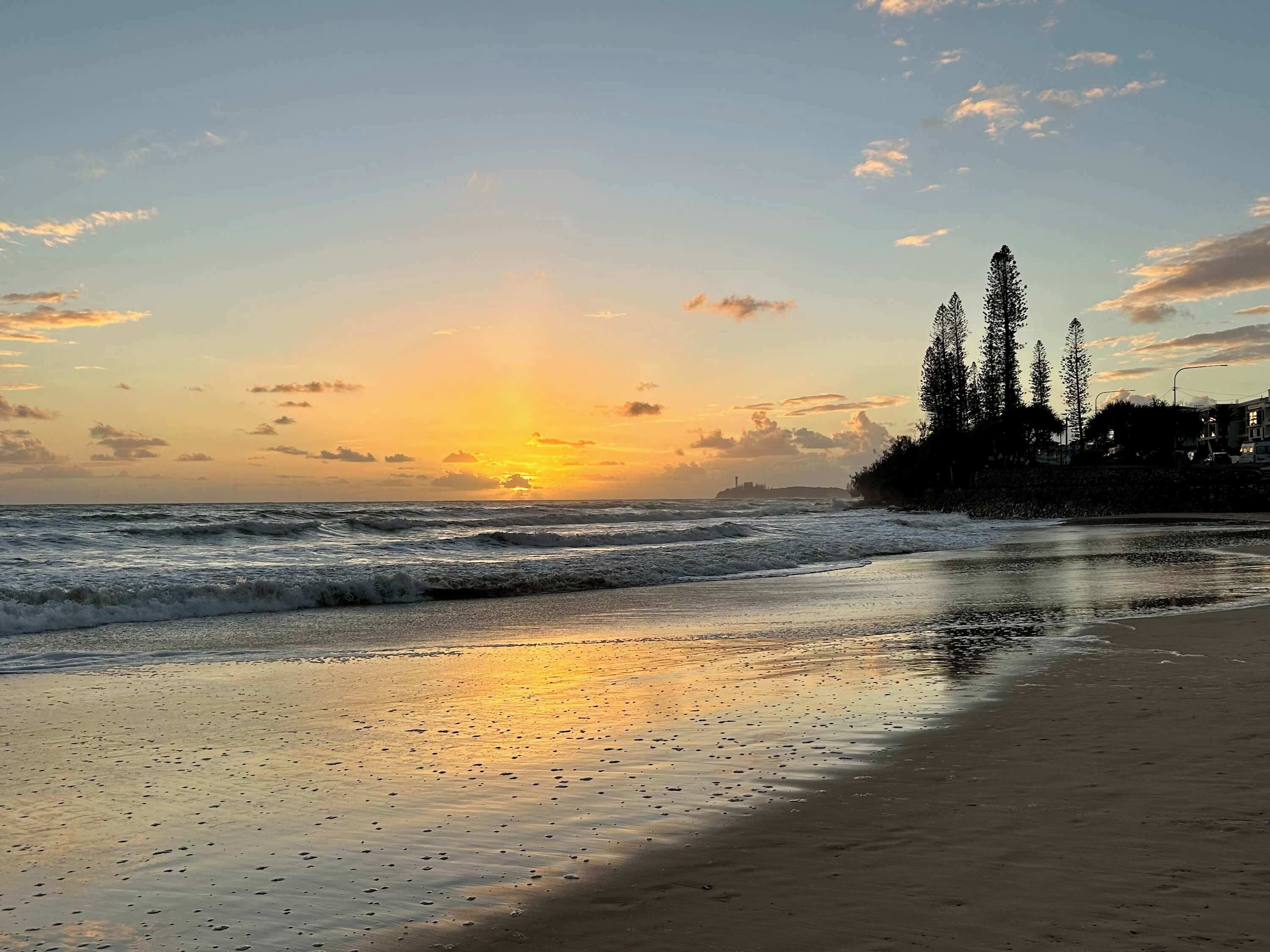 Ein Strand mit Wellen, einem Sonnenuntergang und Bäumen im Hintergrund.