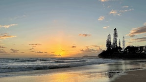 A beach with waves, a sunset, and trees in the background. - Headlands Beachside Gem - 50mtrs To Toes In Sand! (Alexandra Headland)
