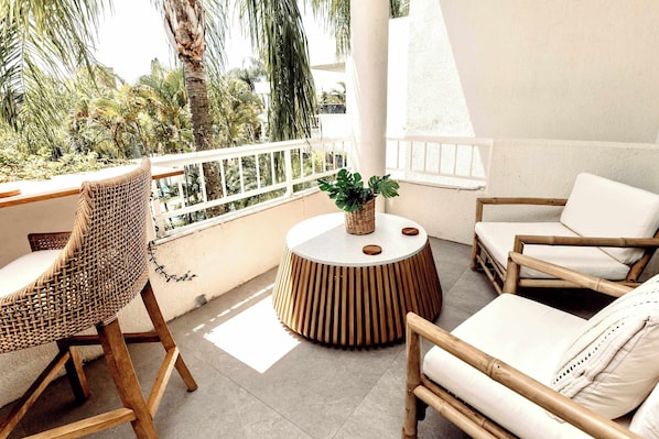 Un balcón con sillas de mimbre, una mesa redonda de madera con una planta y vistas a palmeras.
