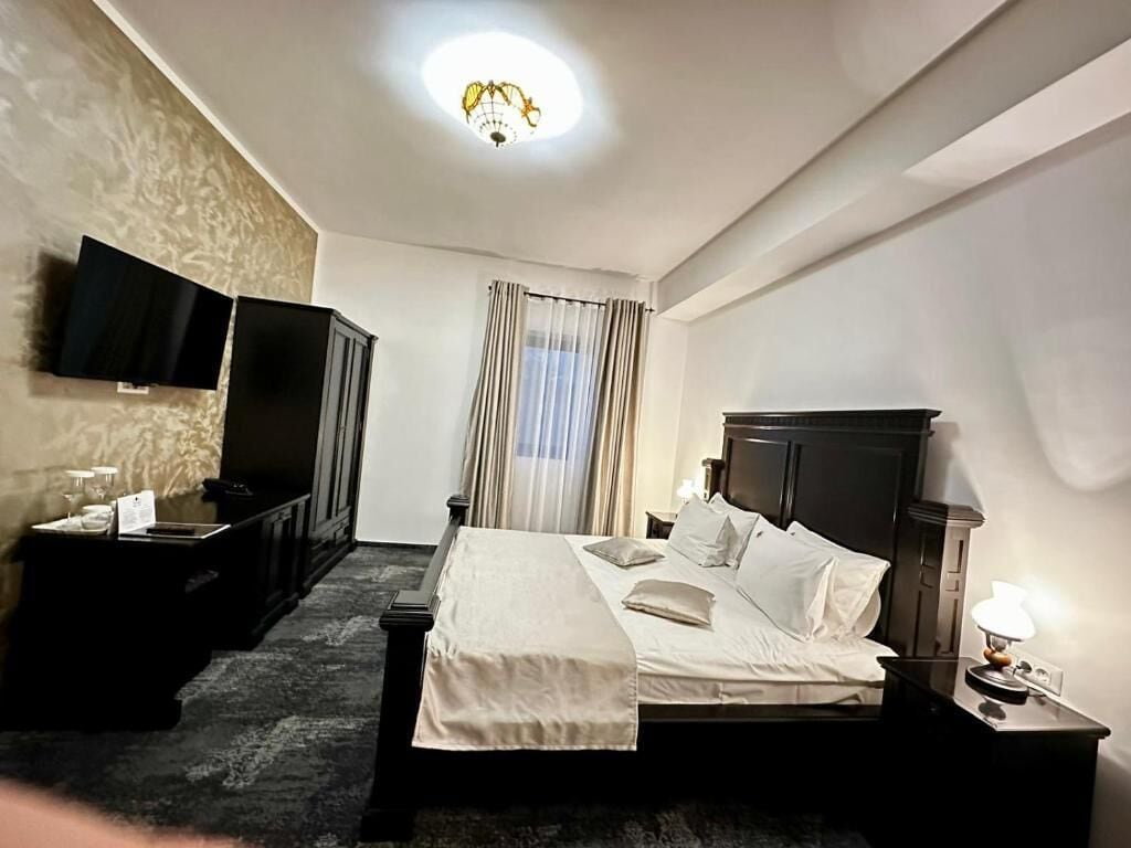 A hotel room with a bed, a TV mounted on the wall, a bedside table with a lamp, and a window with curtains.