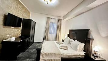 A hotel room with a bed, a TV mounted on the wall, a bedside table with a lamp, and a window with curtains.