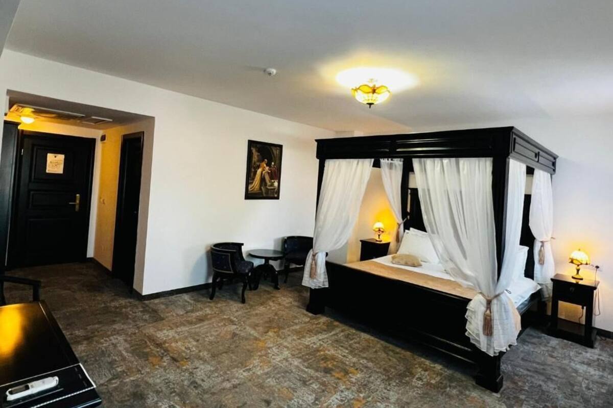 Uma cama com dossel e cortinas brancas, uma escrivaninha com cadeira e uma mesinha de cabeceira com abajur.