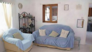 A room with a light blue sofa, a beige armchair, a small table, a window, and a metal shelf with various items. - Good Times in Gingerland, Nevis | 3-Bed Bungalow, Garden & Mountain Views (Gingerland)