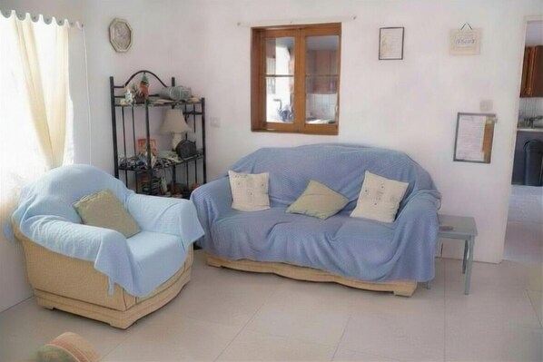 A room with a light blue sofa, a beige armchair, a small table, a window, and a metal shelf with various items.