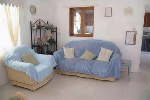 A room with a light blue sofa, a beige armchair, a small table, a window, and a metal shelf with various items.