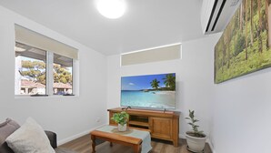 Ein Wohnzimmer mit einem großen Fernseher, einem Holztisch, einer Topfpflanze und Blick auf einen Strand durch das Fenster.