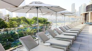 Un área de lounge en la azotea con tumbonas y sombrillas, con vistas a una piscina y al paisaje urbano.