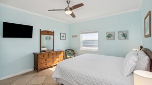 A bedroom with a bed, a dresser, a ceiling fan, a TV, a window, and artwork on the wall.