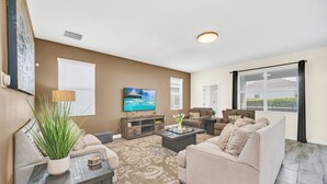 A living room with a sofa, coffee table, and TV. There are windows with blinds, a plant, and a painting on the wall.