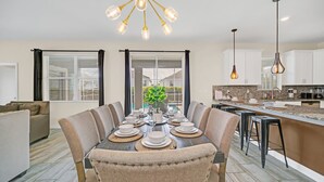 A modern kitchen with a dining area, featuring a large island, pendant lights, and a view to the outside.