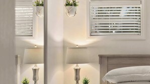 A bedroom with a bed, bedside tables, lamps, a mirror, a window with blinds, and a hanging plant. - PB Bungalow-Private fenced yard-Parking-Remodeled! (San Diego)