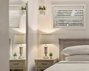A bedroom with a bed, bedside tables, lamps, a mirror, a window with blinds, and a hanging plant.