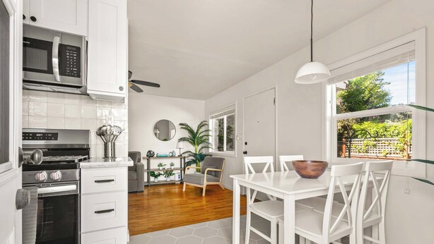 A modern kitchen with white cabinetry, a microwave, stove, and oven. There is a dining area with a white table and chairs, and a large window with a view of greenery. - PB Bungalow-Private fenced yard-Parking-Remodeled! (San Diego)