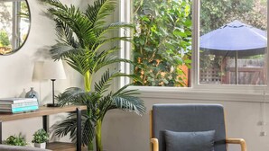 A room with a wooden floor, a grey sofa, a blue armchair, a round mirror, a potted plant, and a window with an umbrella outside. - PB Bungalow-Private fenced yard-Parking-Remodeled! (San Diego)