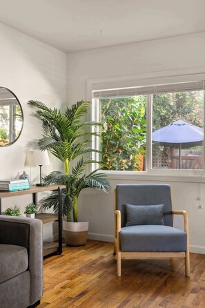 A room with a wooden floor, a grey sofa, a blue armchair, a round mirror, a potted plant, and a window with an umbrella outside.