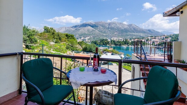 Un balcon avec deux chaises vertes, une petite table sur laquelle se trouve une bouteille de vin, et une vue sur un lac et des montagnes.