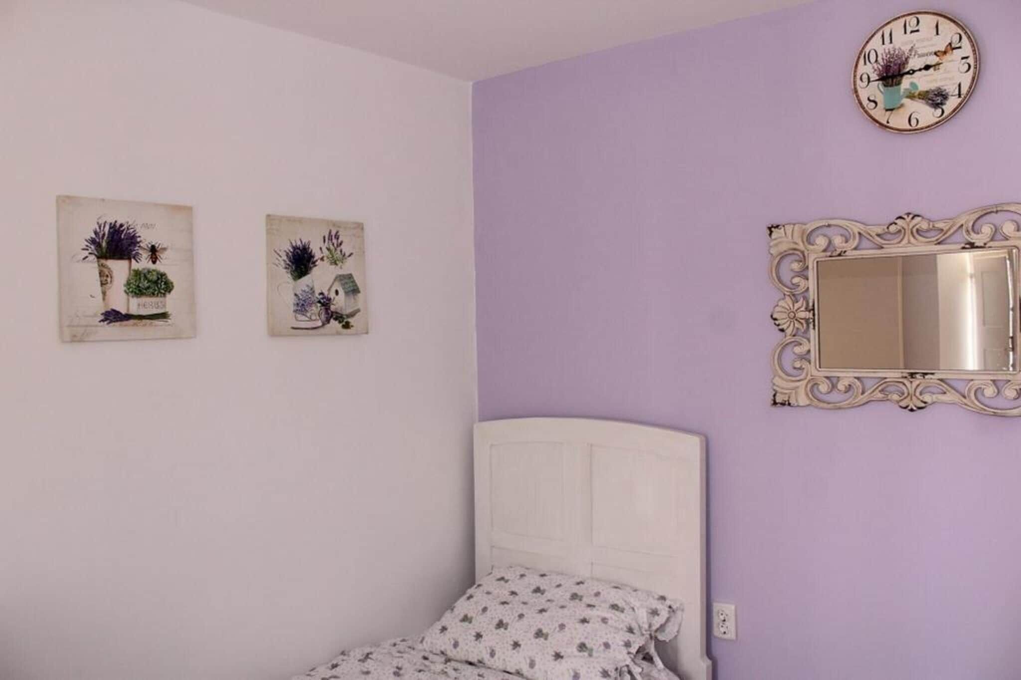 Een slaapkamer met een wit bed, een paarse muur, een decoratieve spiegel en twee kunstwerken met bloemen.