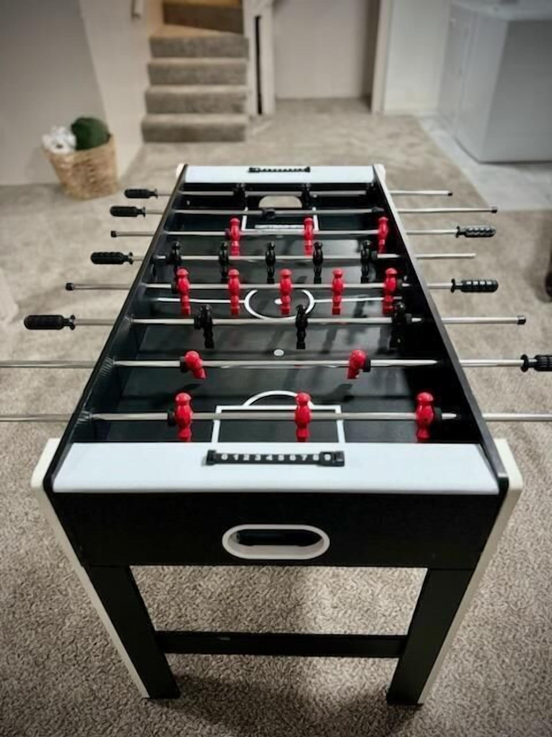 Une table de baby-foot au design noir et blanc, placée dans une pièce dont le sol est recouvert de moquette, avec un escalier en arrière-plan.