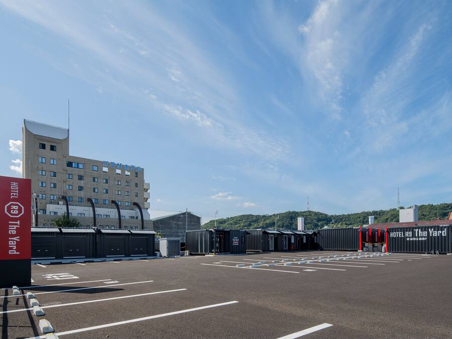 A hotel parking lot with marked spaces, a red sign for HOTEL R9 The Yard, and a large building in the background.
