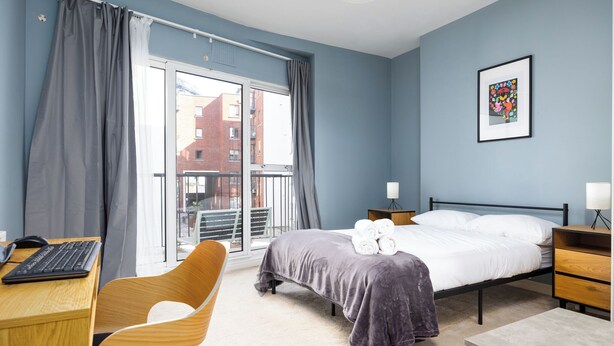 Una camera d'albergo con un letto, una scrivania con un computer, una sedia, un comodino e una finestra con tende.