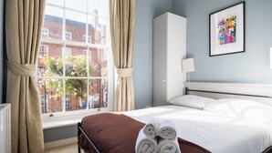Una camera d'albergo con un letto, una finestra con tende, un termosifone, una lampada a parete e un quadro incorniciato.