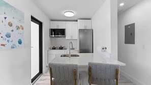 Una cocina moderna con armarios blancos, un refrigerador de acero inoxidable y un microondas. Hay un área de comedor con dos sillas y una mesa.
