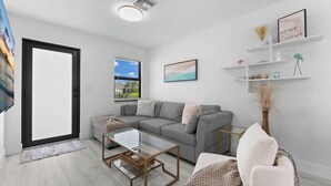 Una sala de estar moderna con un sofá gris, una mesa de centro de vidrio y una pared blanca con un marco de fotos y estantes.