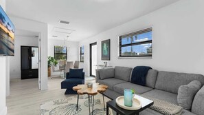 A modern living room with a grey sofa, a blue armchair, and a wooden coffee table. There is a painting on the wall and a window with blinds. - The Ocean @ Naples Boutique (Naples)