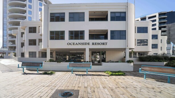 Moderni rakennus, jonka kyltissä lukee ”Oceanside Resort”, ja jonka edessä on puinen kävelyreitti, penkkejä ja kirkas taivas.