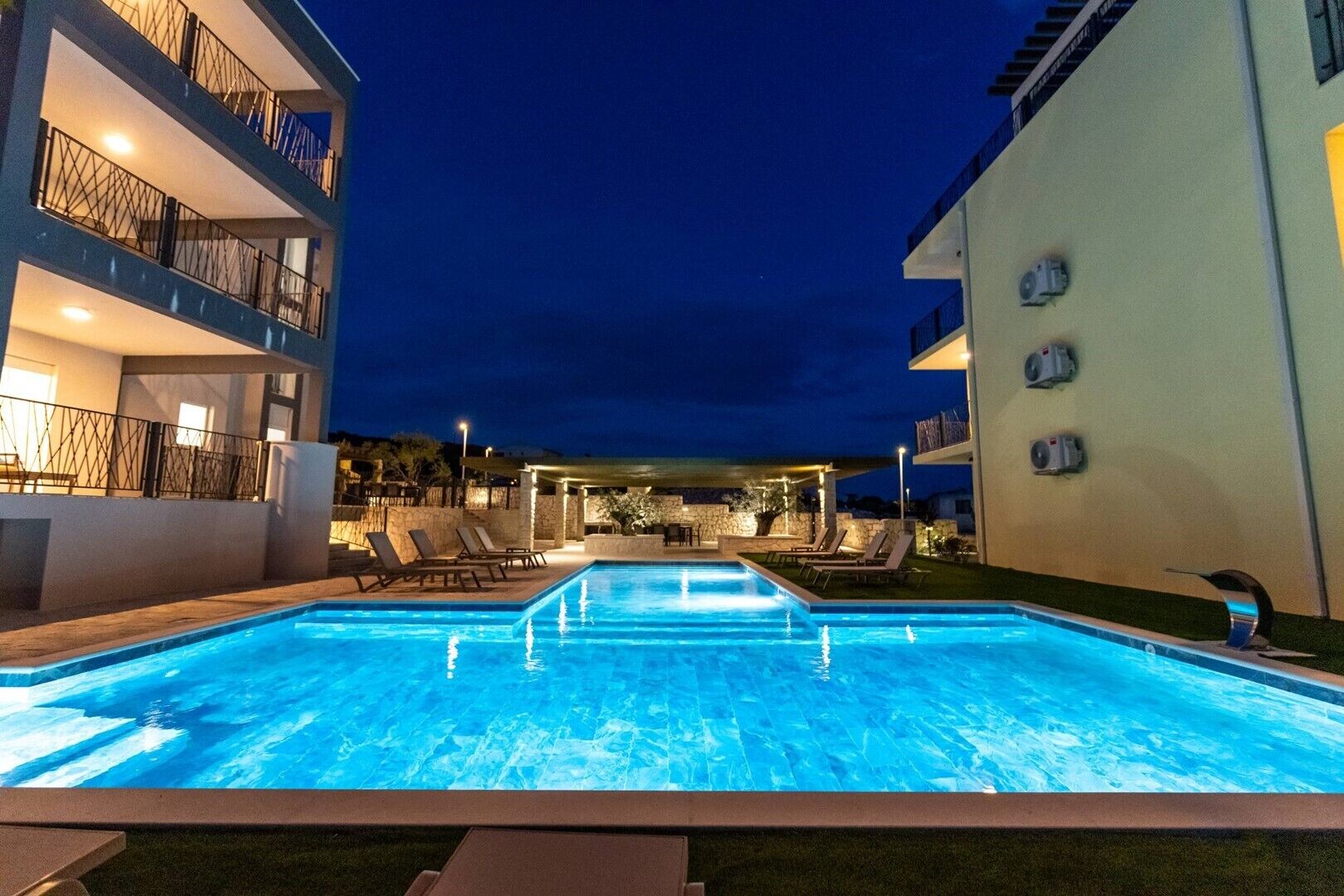 A swimming pool with lounge chairs, a covered seating area, and a building with balconies and air conditioning units.
