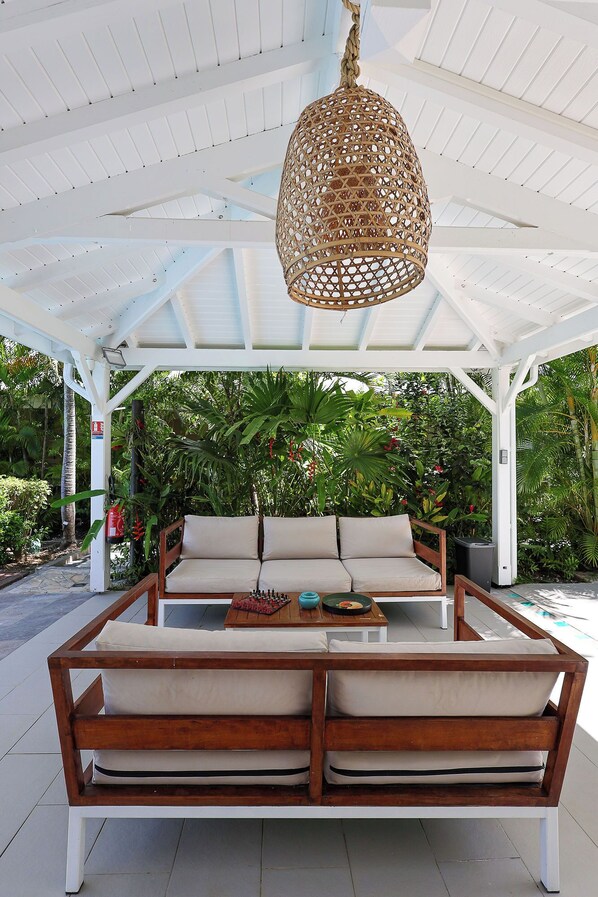 A covered outdoor seating area with a woven hanging light fixture, a cushioned bench with a table, and lush greenery in the background.