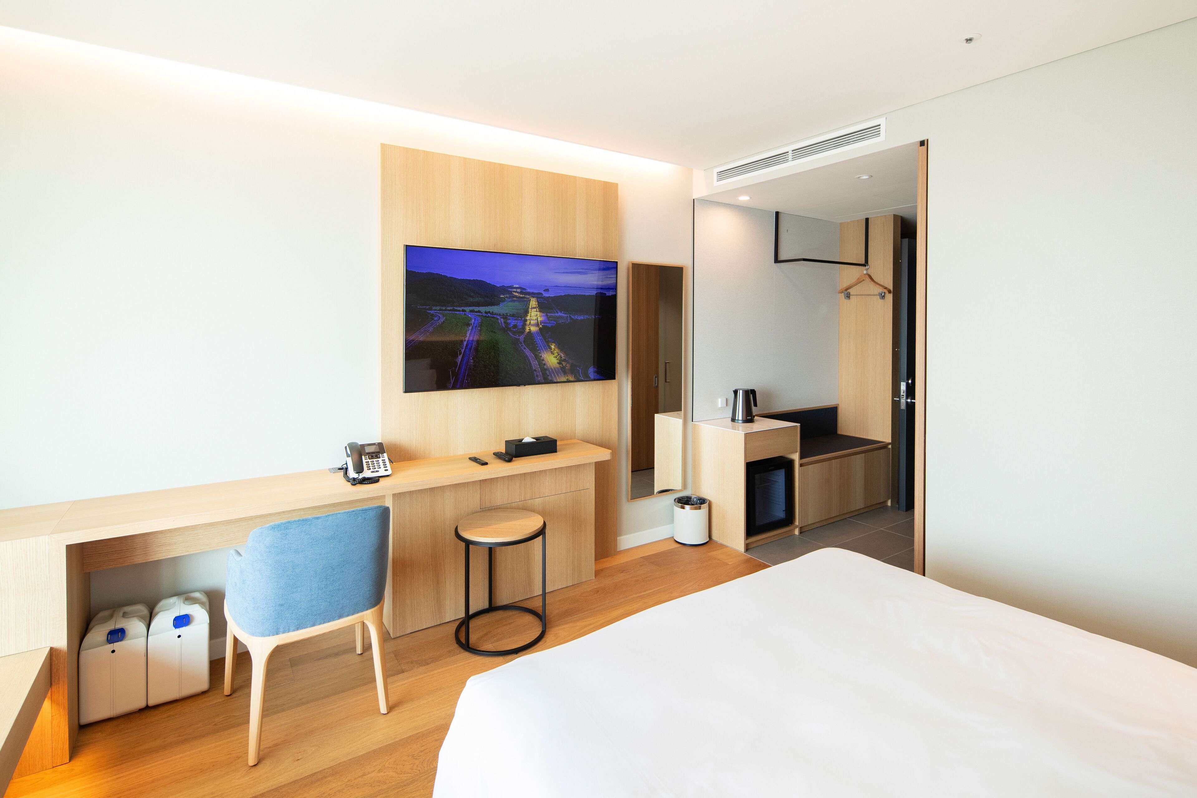 A modern hotel room with a large bed, a desk with a chair, a TV mounted on the wall, and a small kitchenette area.