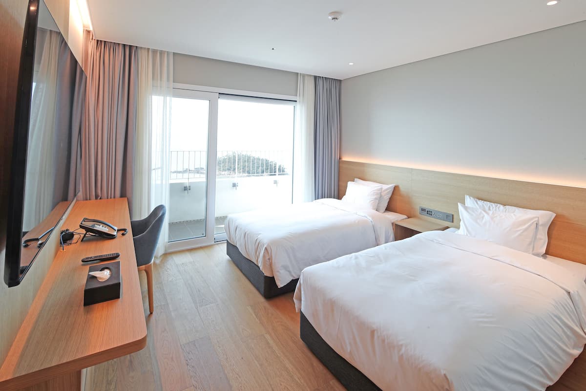Ein Hotelzimmer mit zwei Betten, einem Schreibtisch mit Telefon und Fernbedienung sowie einem Blick auf einen Balkon.