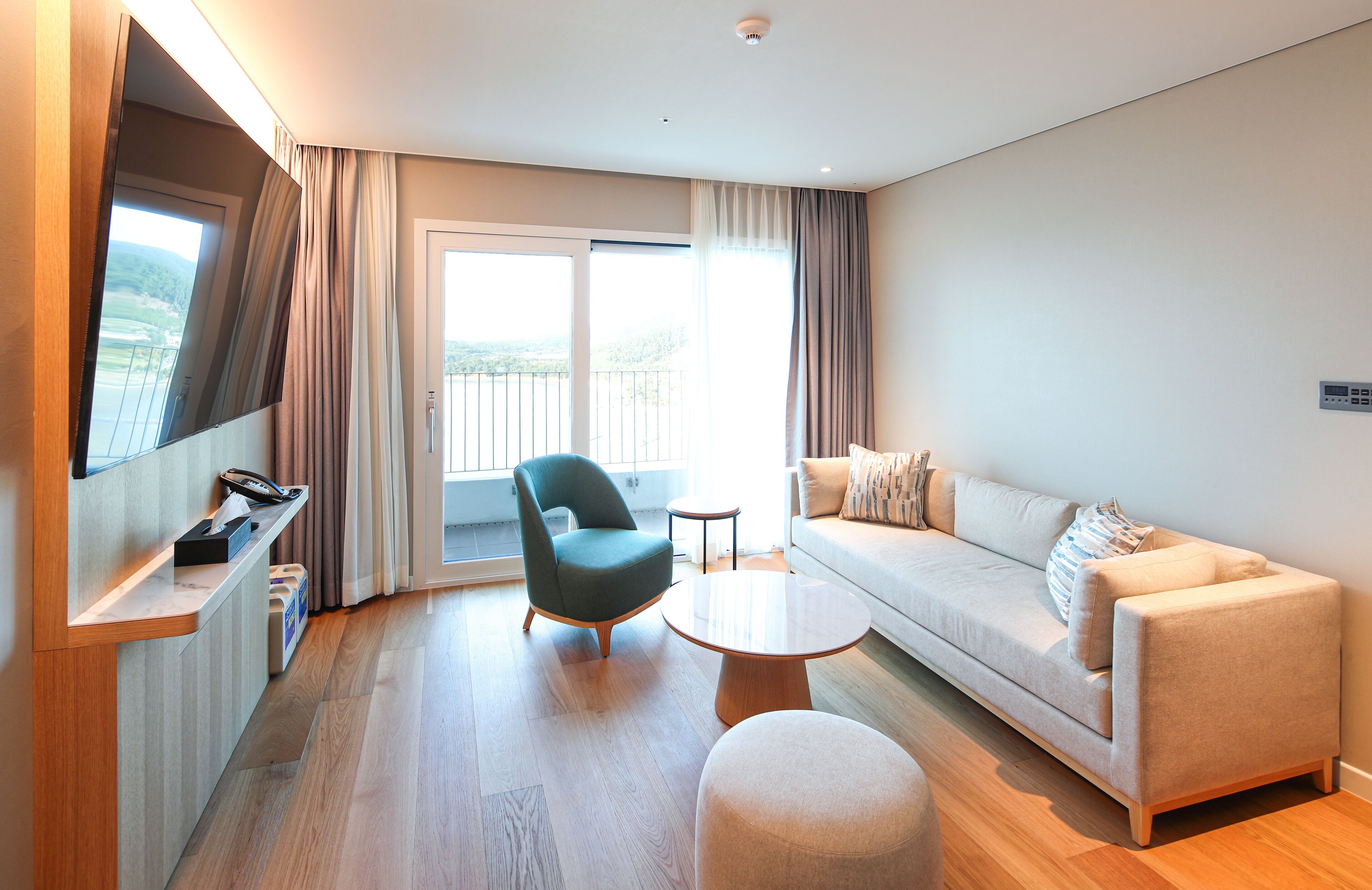 A modern hotel room with a sofa, armchair, and a small table. There is a balcony with a view of the outdoors.