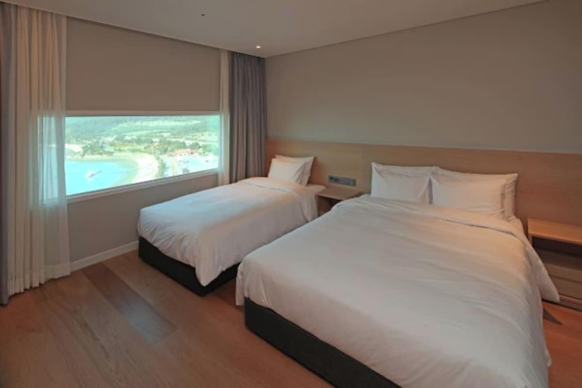 A hotel room with two beds, a large window with a view of a beach, and a wooden floor.