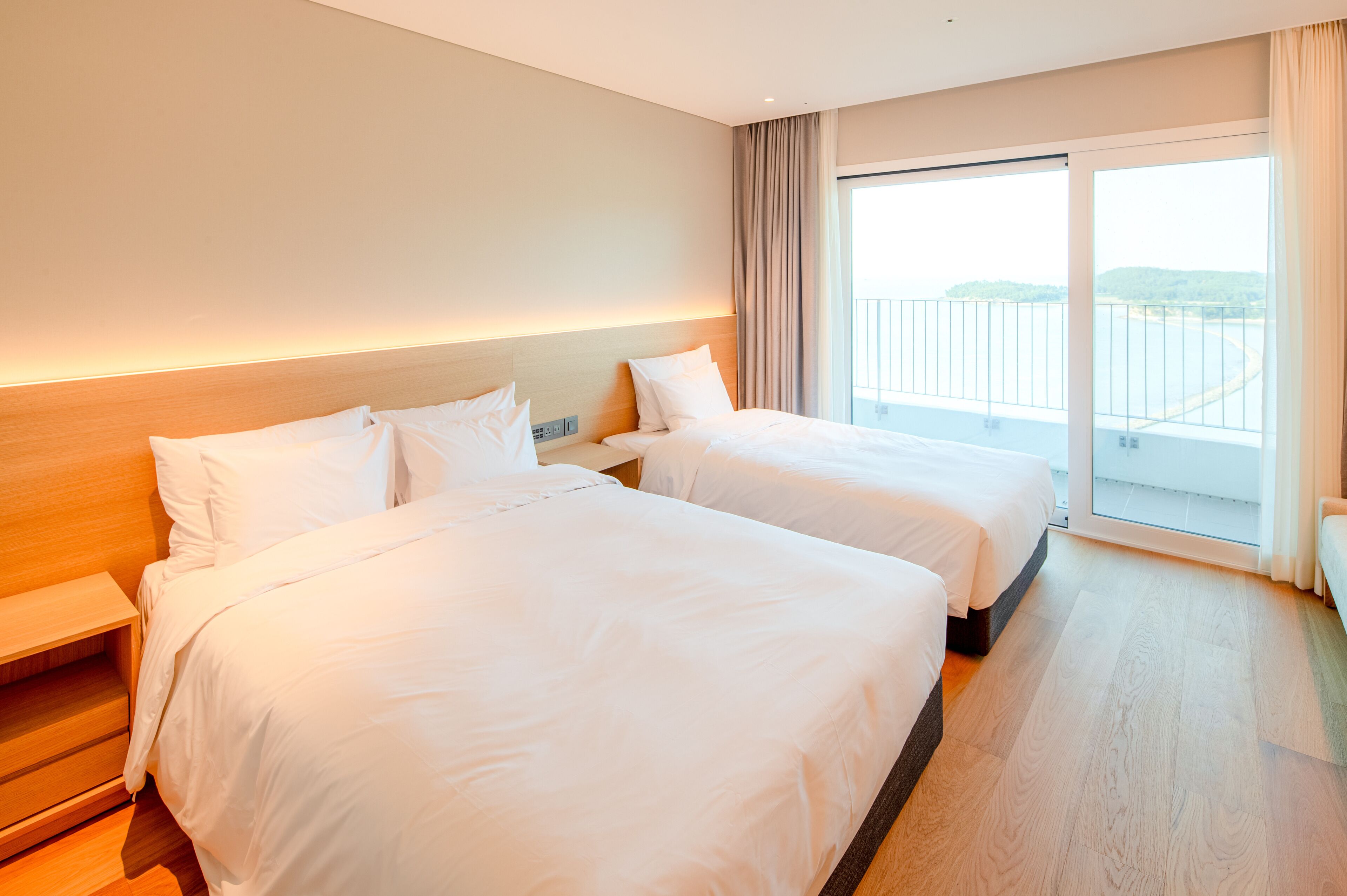 A hotel room with two beds, a wooden headboard, a nightstand, and a large window with a view of the sea.