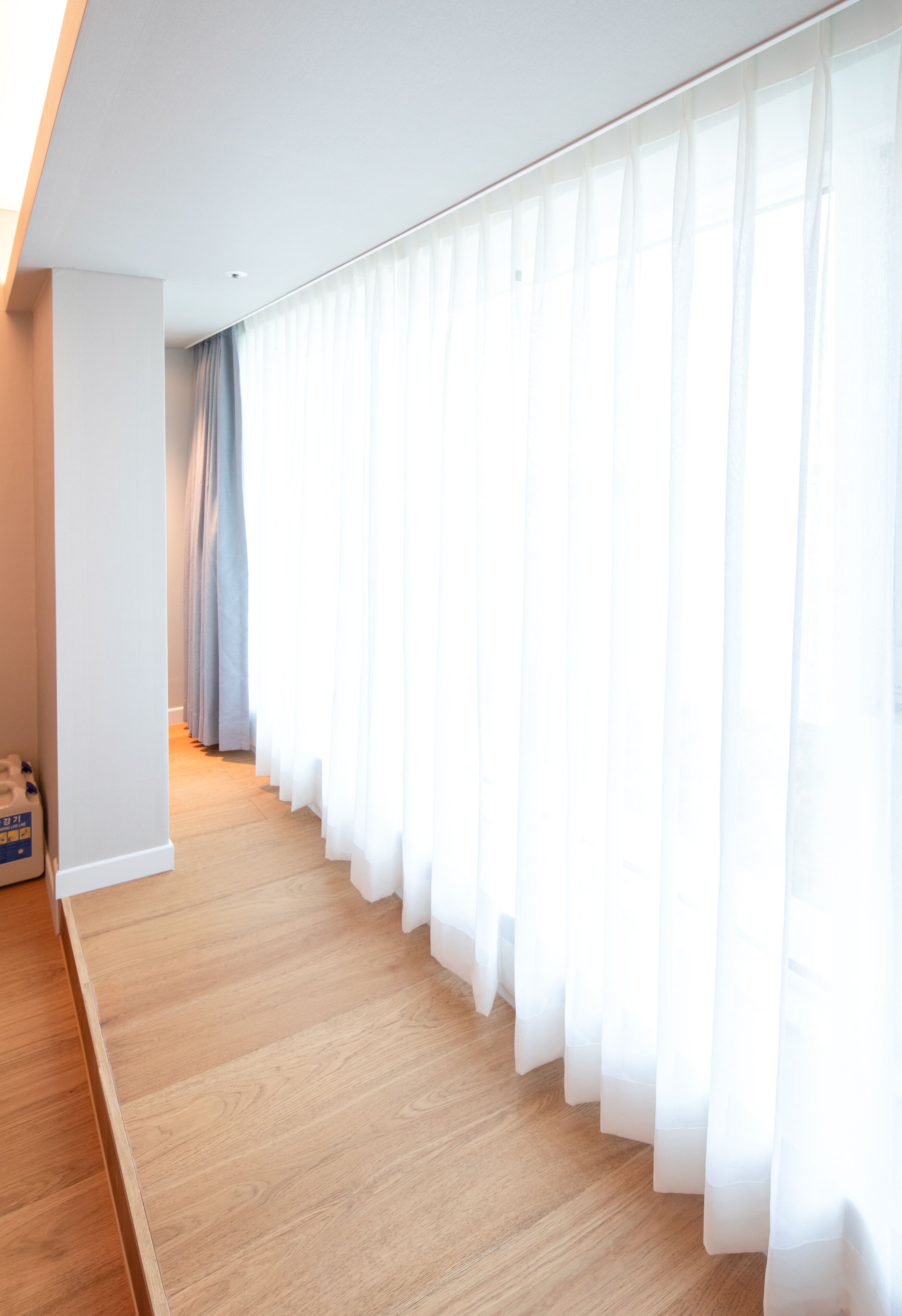 A hallway with wooden flooring, white walls, and sheer white curtains.
