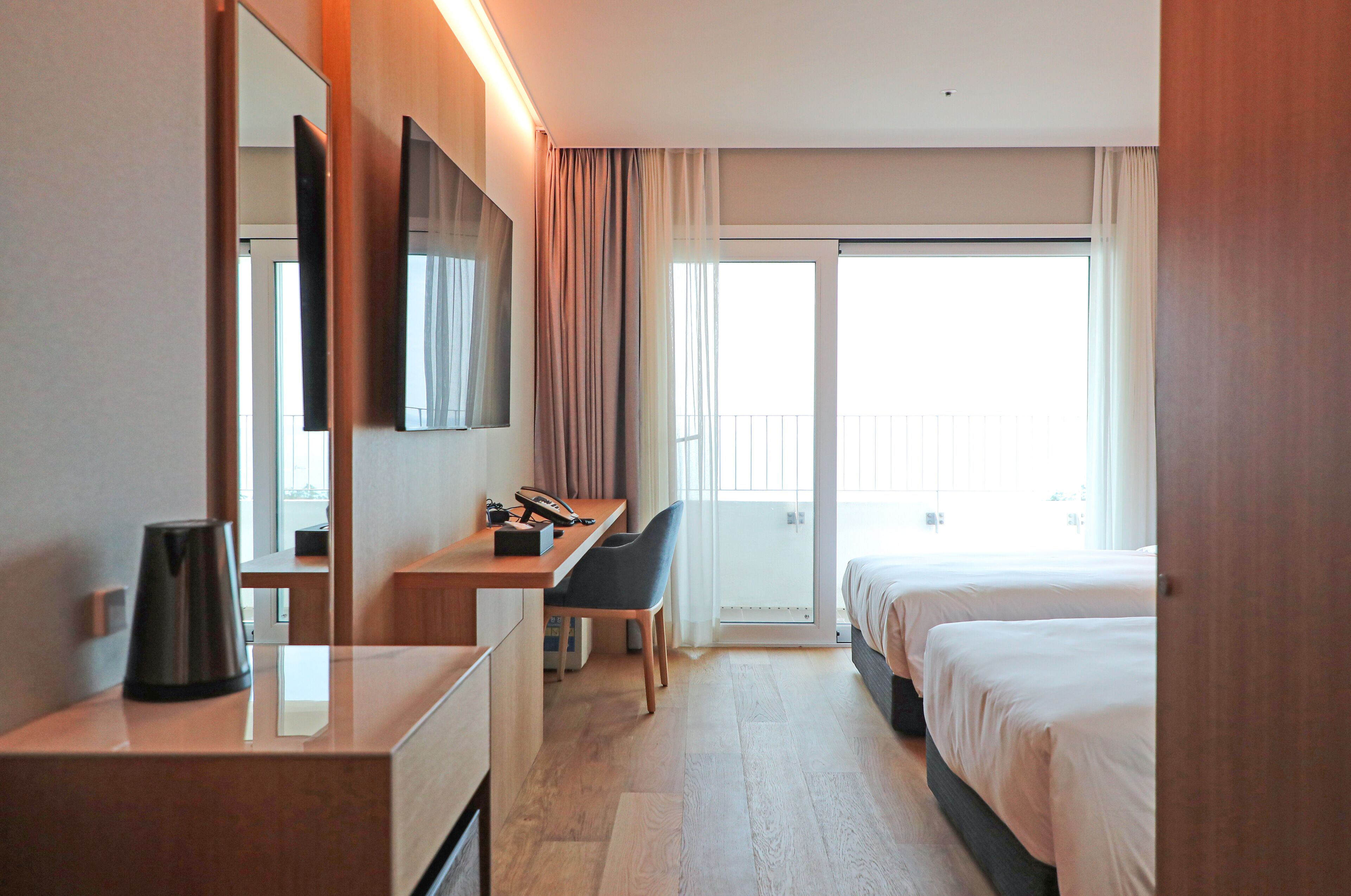 Ein Hotelzimmer mit einem Bett, einem Schreibtisch, einem Stuhl, einem großen Spiegel und Blick ins Freie durch eine Schiebetür.